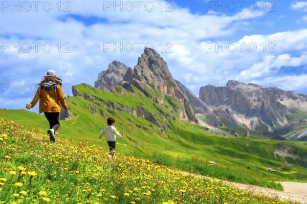 Tourists running through a field of wildflowers with the seceda peaks in the dolomites mountain range of italy in the background on a beautiful summer day
