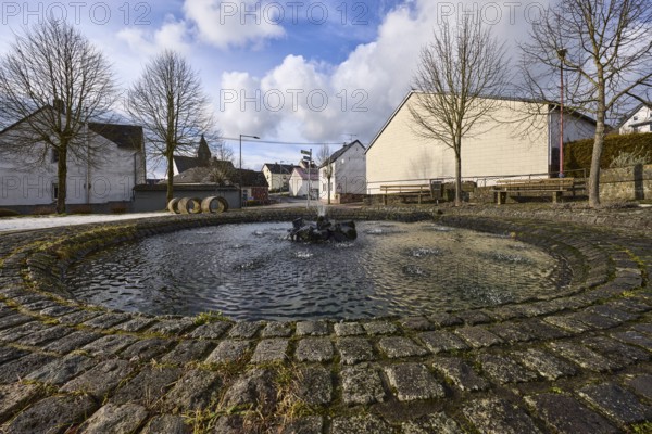 Fountain, water fountains, fountain, houses, trees, blue sky with cumulus clouds, frog perspective, intersection Im Höfchen, Hauptstraße and Heyrother Straße, Dreis-Brück, Eifel, Vulkaneifel district, Rhineland-Palatinate, Germany, Europe