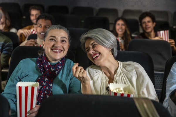 Two cheerful senior women laughing and eating popcorn while watching a movie at the cinema, sharing a joyful moment together