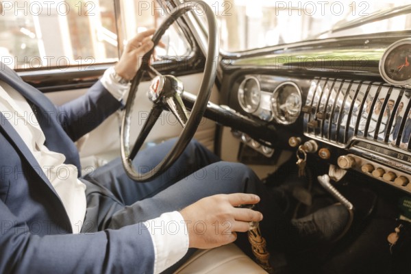 A man in a suit is driving a car with a black steering wheel. The car is a vintage model and the man is wearing a blue suit. Concept of elegance and sophistication