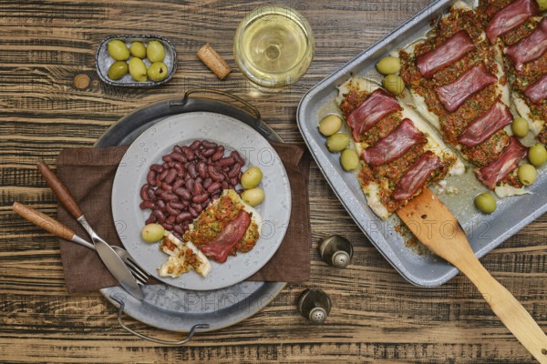 Baked fish topped with a crusty bacon, surrounded by green olives and red beans. A glass of white wine rests nearby, creating a cozy dining atmosphere