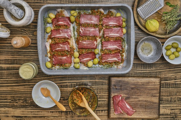 Fresh fish fillets layered with bacon and seasoning, herbs, and olives sits on a baking tray. Surrounding ingredients include spices and utensils, showcasing the artistry of cooking in a home kitchen