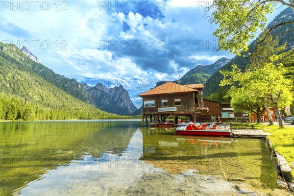 Stunning view of lake dobbiaco with pedal boats and a restaurant, surrounded by the majestic peaks of the dolomites on a sunny summer day