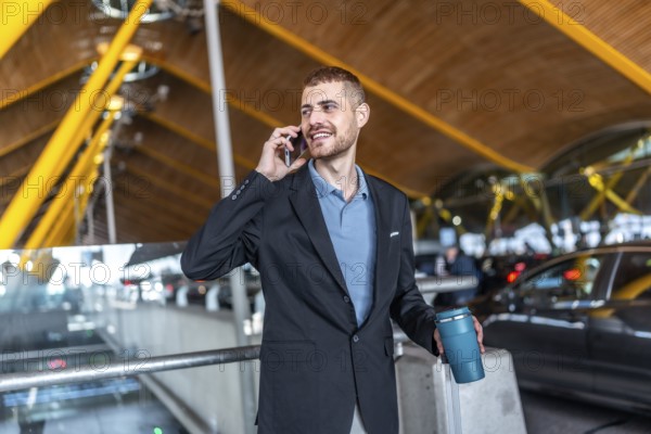 Businessman holding a reusable water bottle and a suitcase while talking on his smartphone at the airport