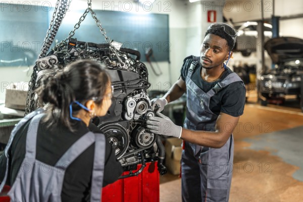Two focused mechanics examining a car engine hanging from a crane in a professional workshop, collaborating on vehicle maintenance