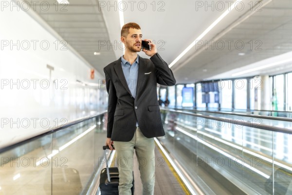 Businessman pulling suitcase and talking on smartphone while walking on moving walkway in airport