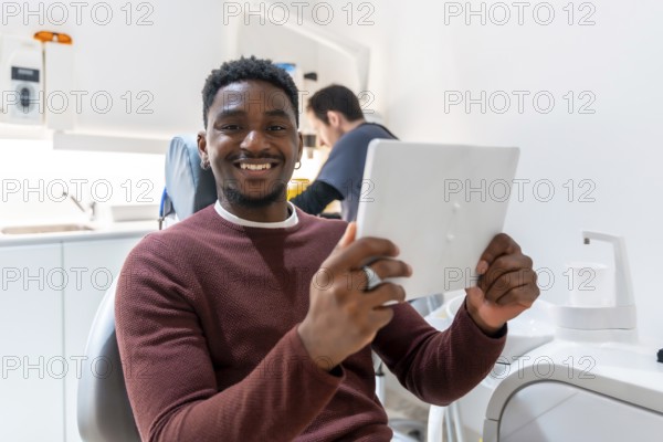 Smiling patient using digital tablet, possibly reviewing treatment plan or x rays, while dentist works in background of modern dental clinic