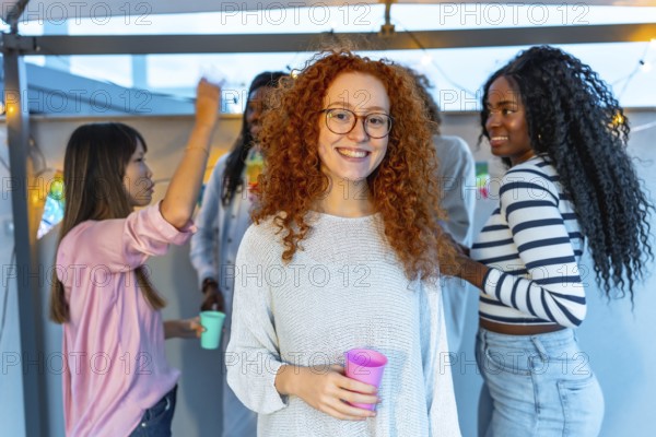 Young woman with redhead curly hair and eyeglasses smiling standing with beer glass in a roof party with multi-ethnic friends