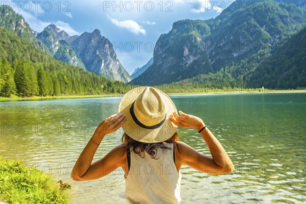Young woman holding her straw hat admiring the landscape of lake dobbiaco and the dolomites during summer vacation in italy