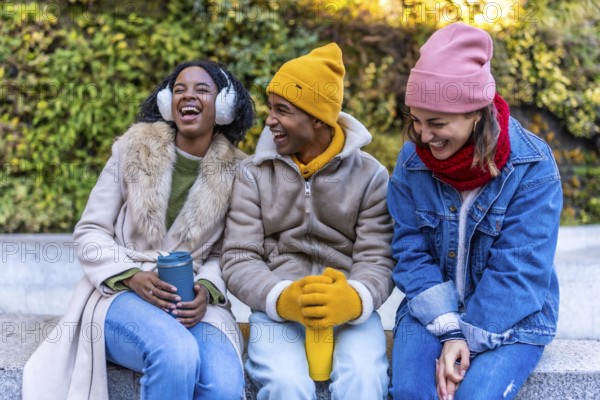 Three young multi ethnic friends, two women and a man, are sitting outdoor in winter clothes, laughing and having fun