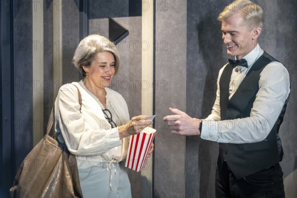 Elegant mature woman handing ticket to smiling usher while holding popcorn bucket, ready to enjoy movie session