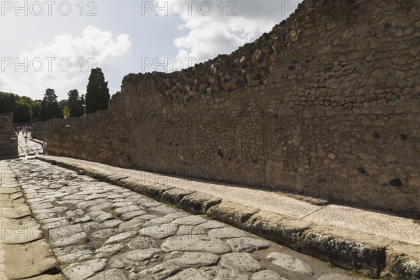 Long narrow stone sidewalk and street made of large grey smooth and worn lava stones plus remnant of old stone and terracotta brick wall structure at the ancient ruins of Pompeii, Campania region, Italy, Europe