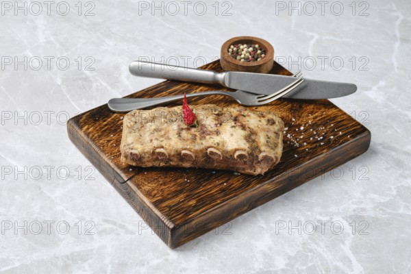 Roasted pork ribs on a rustic wooden board, accompanied by a knife and fork. A small bowl of mixed pepper serves as an elegant touch, enhancing the dining experience