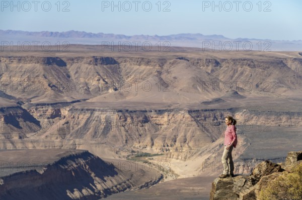 Woman standing on a rock at the edge of the precipice in front of the gorge landscape of the Fish River Canyon, Fish River Gorge, Namibia, Africa