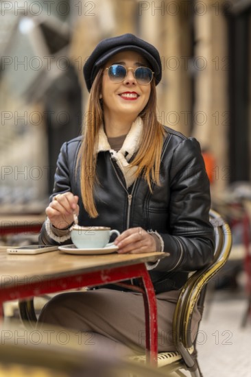 Young woman wearing sunglasses and baker boy hat stirring cappuccino at outdoor cafe table with smartphone nearby