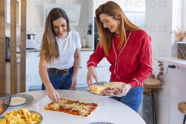 Two happy women adding cheese to pizza dough, preparing a meal with tacos and tortilla chips in a modern kitchen