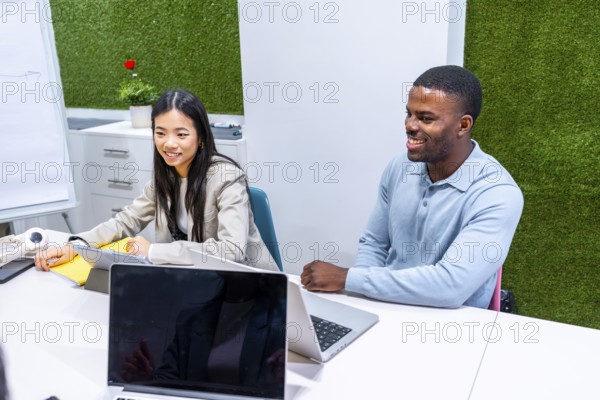 Two businesspeople are working together on a project in a modern coworking office, using laptops and tablets, with a green wall in the background