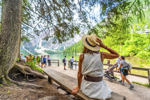 Woman wearing a straw hat relaxing on a bench, enjoying the landscape of lake braies, a famous tourist destination in the dolomites, italy
