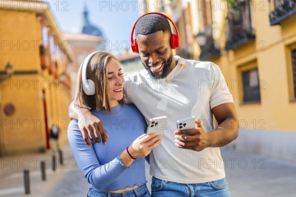 Happy multi-ethnic couple walking embraced and listening music using mobile in the city
