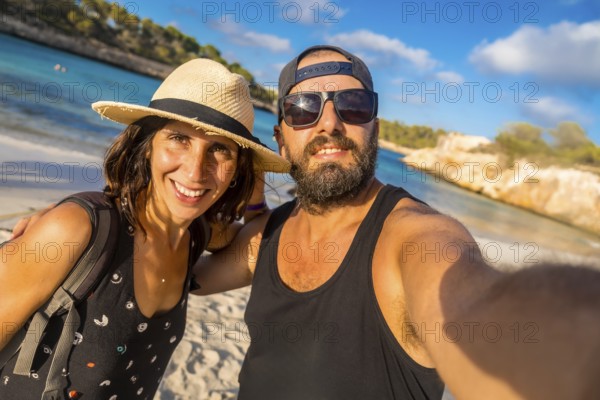A man and woman are posing for a picture on a beach. The man is wearing a black tank top and sunglasses, while the woman is wearing a straw hat. They both have smiles on their faces