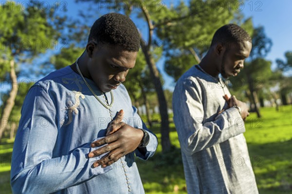 Two young senegalese men wearing traditional dashiki clothing are praying outdoors in a park, expressing their faith and devotion