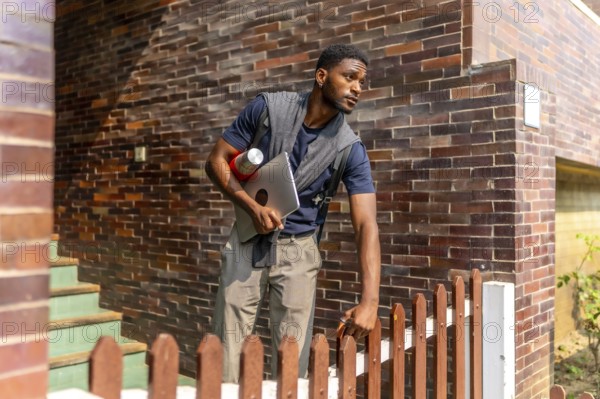Young african businessman leaving a house in a residential district to go to work holding a laptop and a coffee thermos