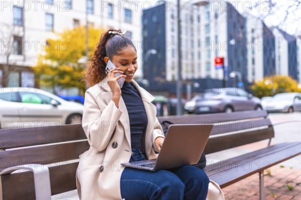African businesswoman talking to the phone and using laptop sitting on a city bench