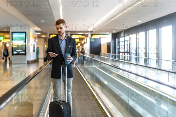 Businessman navigating airport terminal, using smartphone and enjoying coffee while walking on moving walkway
