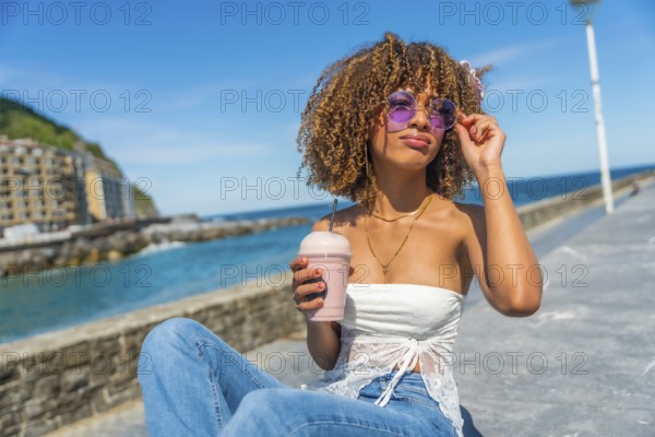 Cool woman with curly hair drinking take away iced juice next to the sea