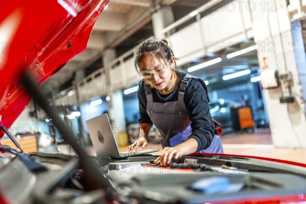Young woman working as a mechanic, using a laptop while inspecting the engine of a red car in a busy garage workshop