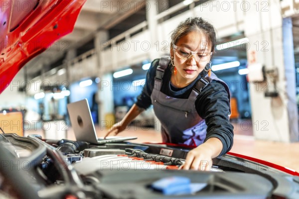 Young woman wearing mechanic overalls inspecting car engine using a laptop in a modern workshop