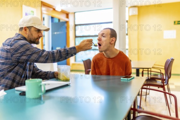 Man helping to eat to a disabled man in a cowokring cafeteria