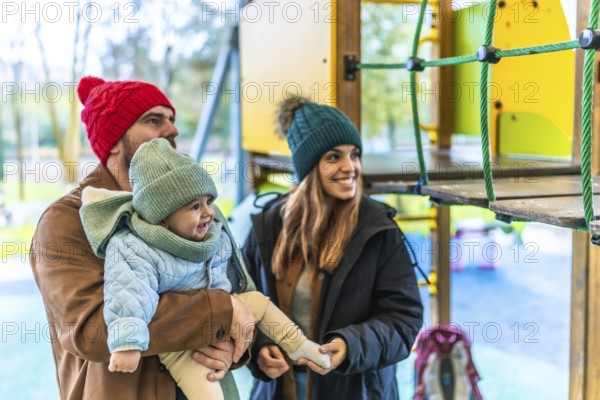Smiling parents holding their baby at the playground, wearing winter clothes, enjoying a family moment