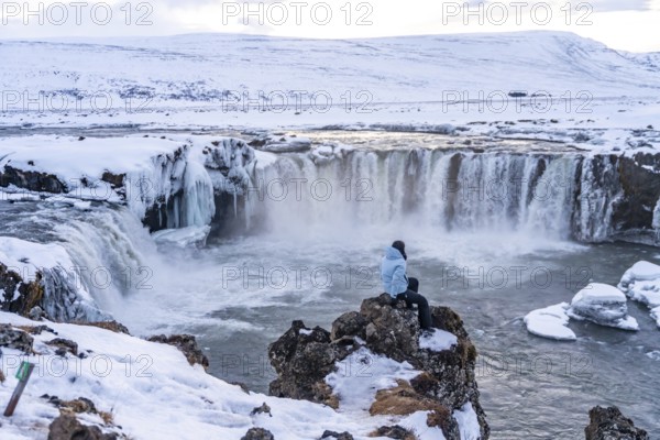 Woman hiker sitting looking at frozen and snowy Godafoss waterfall in winter in Iceland