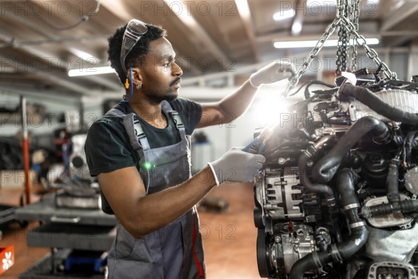 Young mechanic wearing protective gear working on a car engine suspended by a chain hoist in a professional workshop