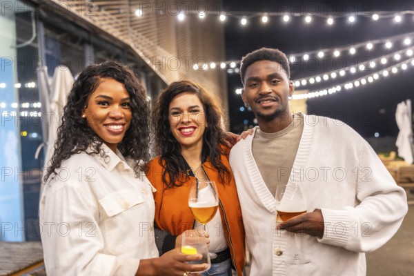Portrait of three multi-ethnic friends enjoying night at bar drinking beer and smiling at camera