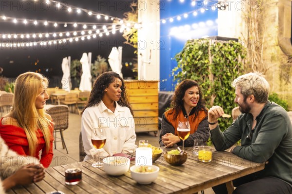 Multi-ethnic male and females friends talking relaxed drinking alcohol and eating snacks in a night terrace bar