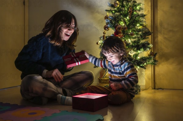 A woman is giving a child a red present. The child is sitting on the floor and the woman is kneeling down. Scene is joyful and festive, as it is a Christmas scene