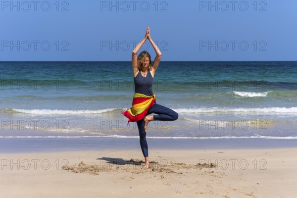 A woman doing yoga on a sandy beach by the sea. Vrksasana or tree. Treen Cove, Cornwall, England, Great Britain
