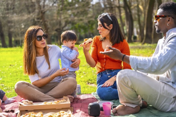 Family and friends enjoying pizza while picnicking in a vibrant green park, laughing and sharing joyful moments under the sunny sky