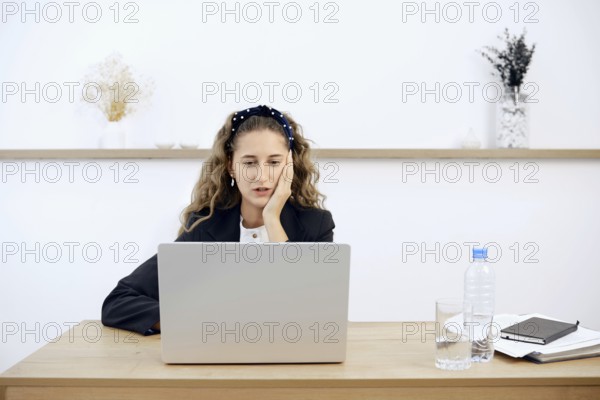 A young woman sits at a wooden desk, focused on her laptop while resting her chin on her hand. A water bottle and glass sit nearby, alongside notebooks and a notepad, in a bright, airy office.