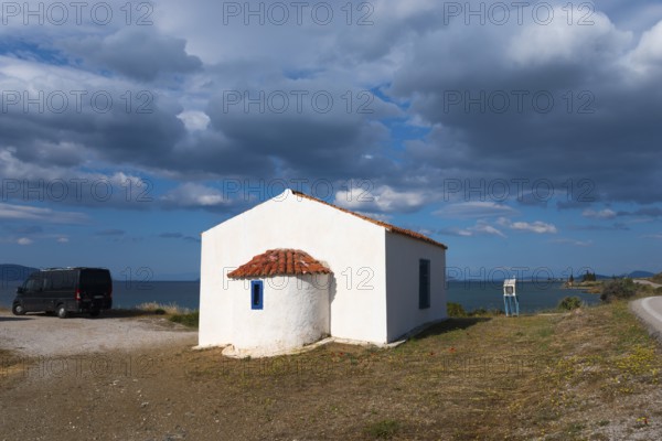White building with a red roof by the sea, surrounded by a cloudy sky and peaceful countryside. A motorhome is parked next to it. Chapel near Vlacheika, Vlachaiika, Attica, Peloponnese, Greece, Europe