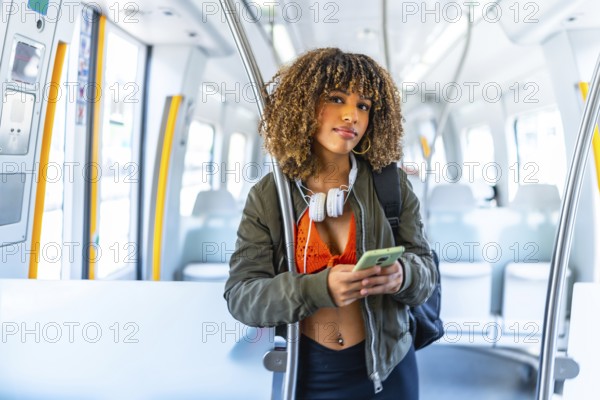 Latin woman with curly hair standing on the train using cellphone and looking at camera