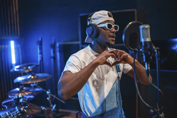 Young stylish male singer making heart shape with hands while recording new hit song in professional music studio