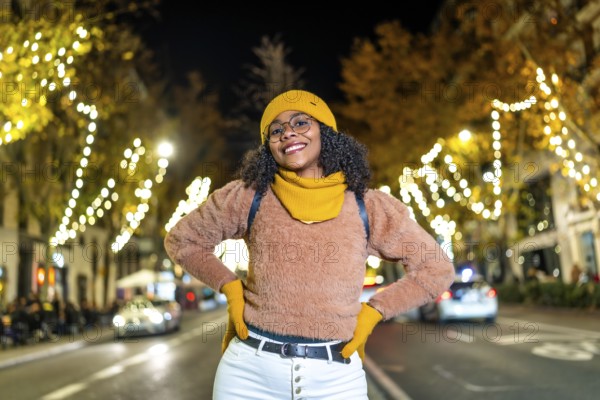 Young black woman tourist wearing yellow winter clothes and backpack, smiling with hands on her hips in a decorated city street at night