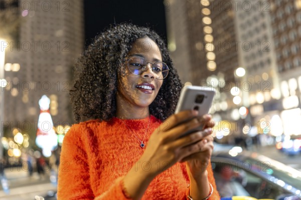 Young woman wearing an orange sweater, smiling while using a smartphone on a bustling city street, surrounded by festive holiday lights at night