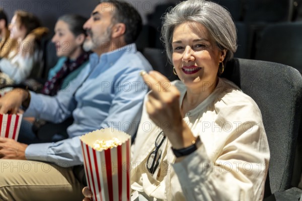 Mature woman smiling and eating popcorn while watching a movie at the cinema with friends, enjoying a leisure activity