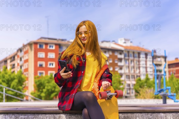 Skater woman in a yellow t-shirt, red plaid shirt and sunglasses, sitting with skateboard in the city, smiling calling with the phone