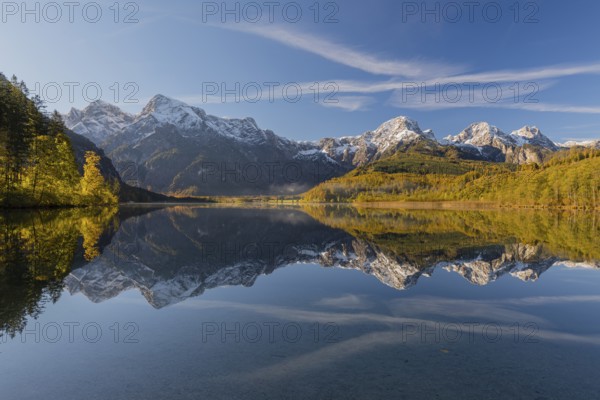 Lake Almsee in autumn with a forest and mountains reflected in the lake. A slowly dissipating fog moves over the lake. Blue sky