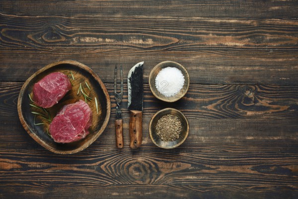 Two raw steaks on a wooden plate alongside bowls of salt and pepper, with old forged knife and fork placed next to them, a cooking session or barbecue preparation
